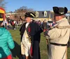 Two people dressed in historical military costumes are participating in an outdoor community event with a crowd that includes children playing