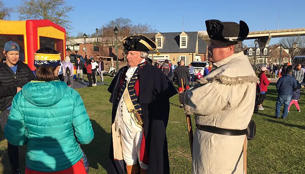 Two people dressed in historical military costumes are participating in an outdoor community event with a crowd that includes children playing