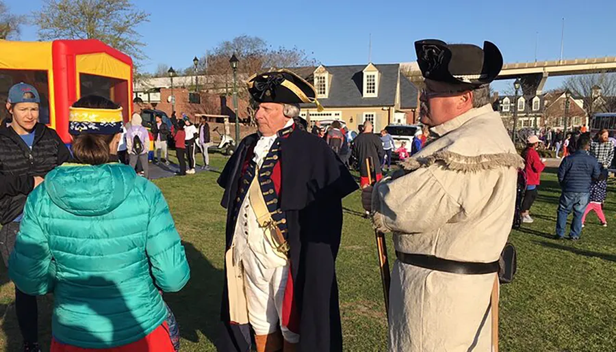 Two people dressed in historical military costumes are participating in an outdoor community event with a crowd that includes children playing.