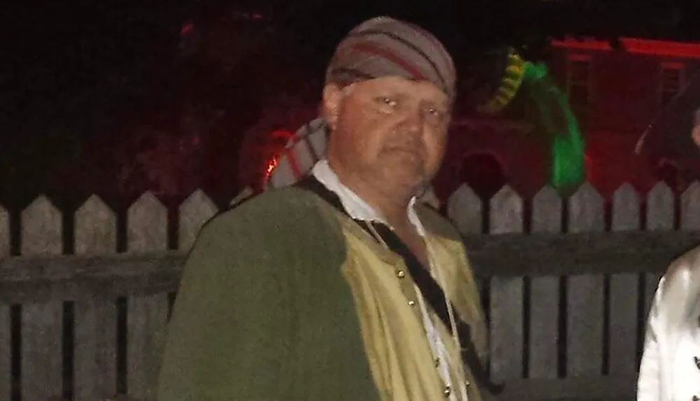 A person with a solemn expression is wearing a beanie and a jacket standing at night with a white picket fence and subtle party decorations in the background