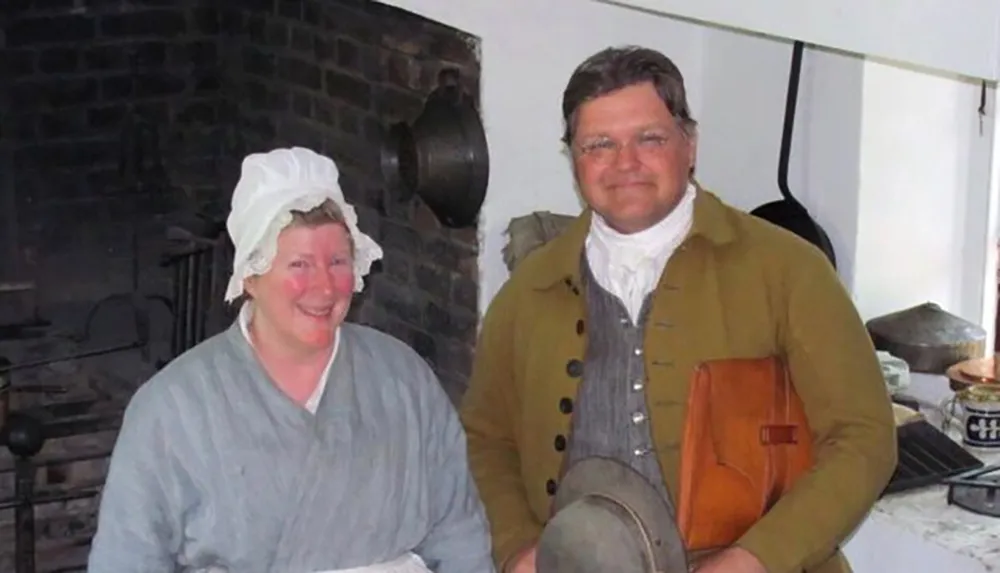 Two individuals dressed in historical clothing are posing with a smile in what appears to be a period-appropriate kitchen setting