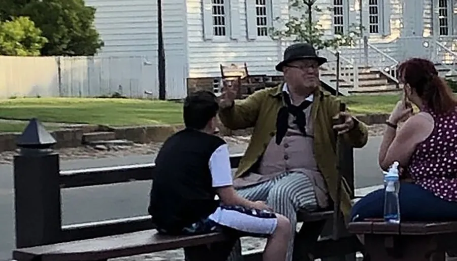 A person dressed in historical costume is gesturing while talking to two people seated on a park bench.