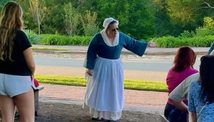 A person dressed in historical clothing is gesturing while speaking to a group of people outdoors.