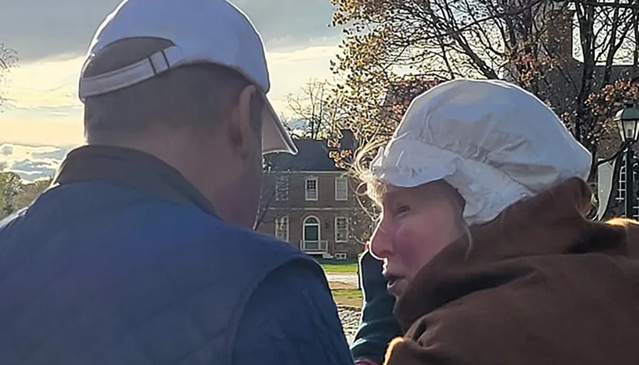 Two people wearing caps are engaged in a close conversation, with one in historical attire, backdropped by a sunlit colonial style building and trees.