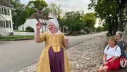 A person dressed in historical costume is gesturing while speaking to onlookers on a cobblestone street lined with colonial-style houses.