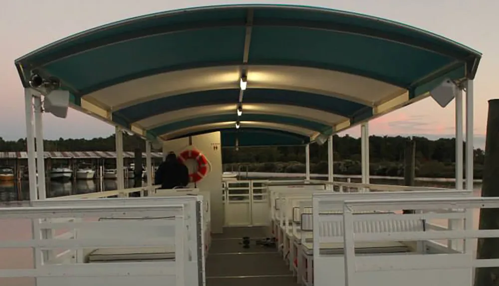 The image shows the inside of a covered open-air boat with rows of white benches docked near a marina at sunset