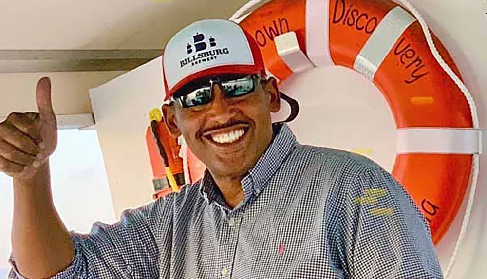 A smiling man in sunglasses and a cap gives a thumbs-up standing in front of an orange lifebuoy on a boat