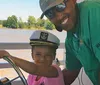 A man and a child both smiling are steering a boat with the child wearing a captains hat