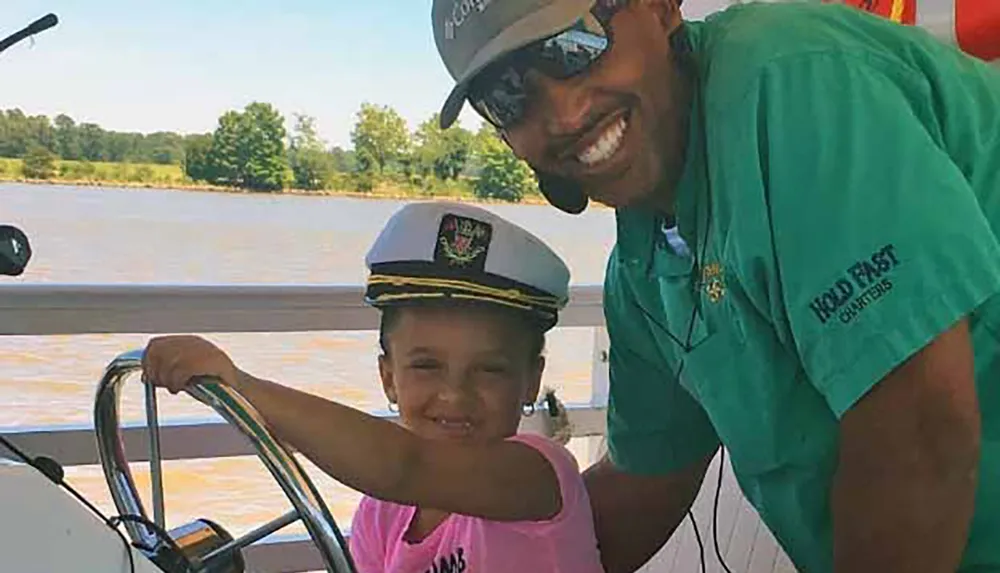 A man and a child both smiling are steering a boat with the child wearing a captains hat