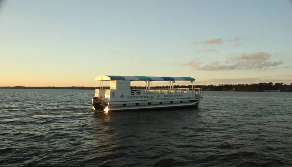 A small tour boat called Jamestown Discovery is cruising on calm waters under a clear sky during sunset