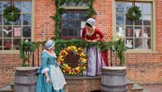 Two women in period costumes decorate a festive Christmas wreath on a brick building's balcony, while another woman watches from inside.