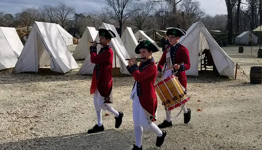 Individuals dressed in historical military uniforms march with a fife and drums in a camp setting with canvas tents.