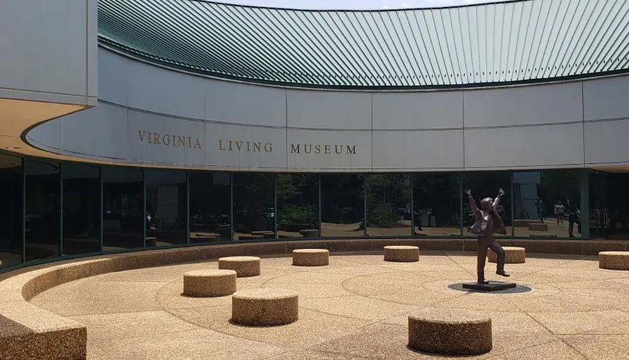 The image shows the exterior of the Virginia Living Museum with its name on the facade and a bronze statue in the courtyard.