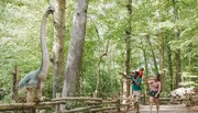 A family is interacting with a life-size dinosaur model in a forested outdoor setting.