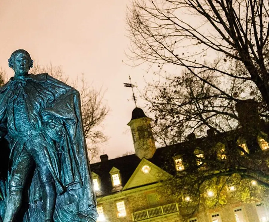 An illuminated bronze statue stands in the foreground with a historic building and a leafless tree in the background against a dusky sky.