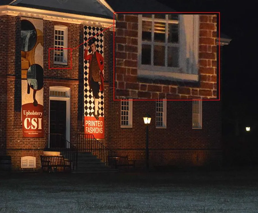 This image shows a nighttime view of a brick building with illuminated windows and posters on the exterior wall, with one window zoomed in on in a red inset, possibly suggesting something of interest there.