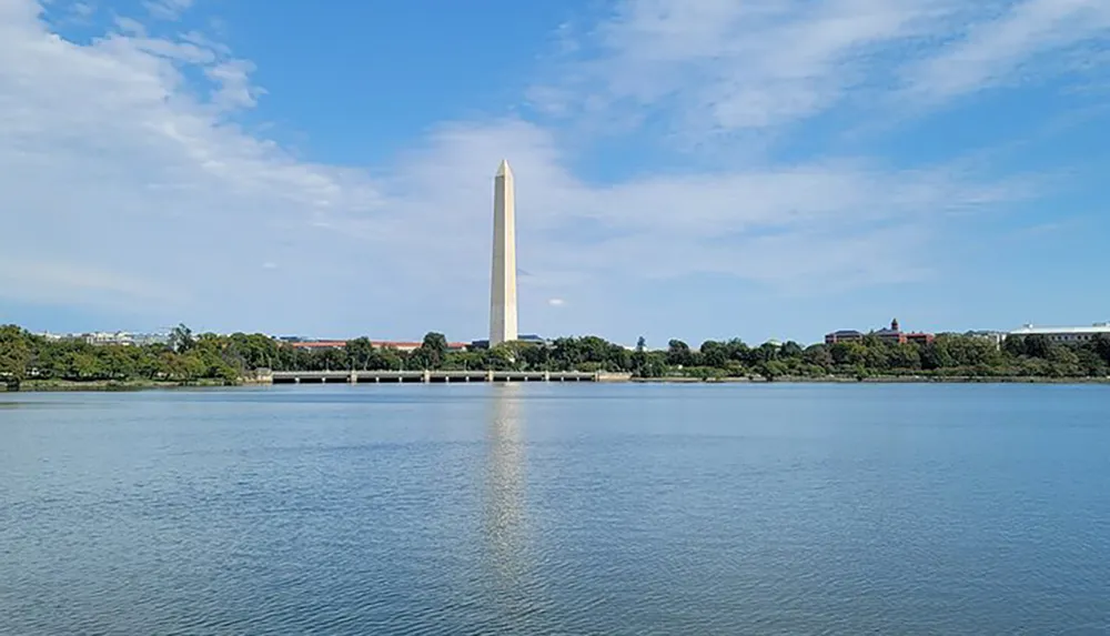 The image shows a daytime view of the Washington Monument towering over the Tidal Basin in Washington DC with a clear sky and calm waters reflecting the monument