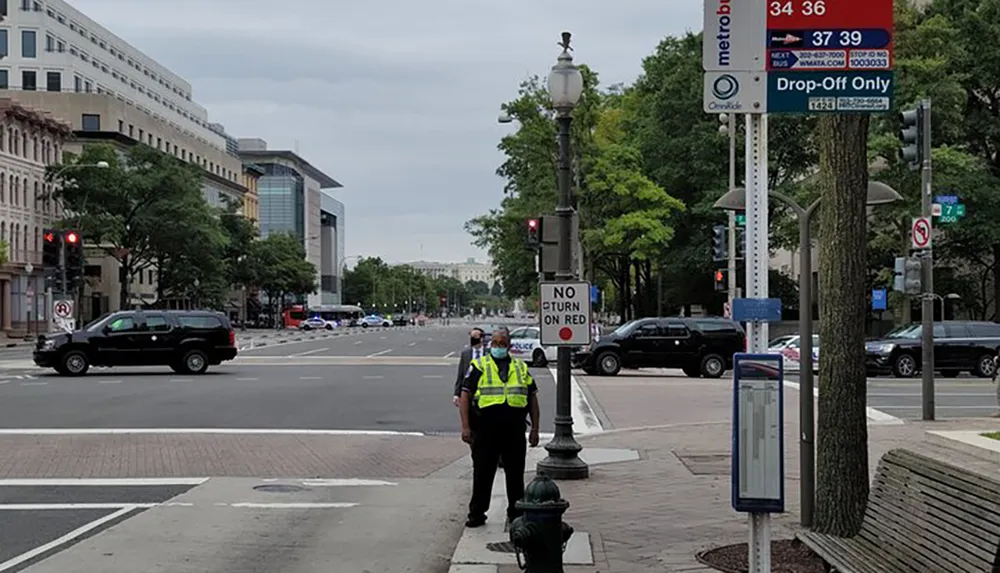 A security officer in a hi-vis vest stands at a street corner in an urban environment with traffic and buildings in the background