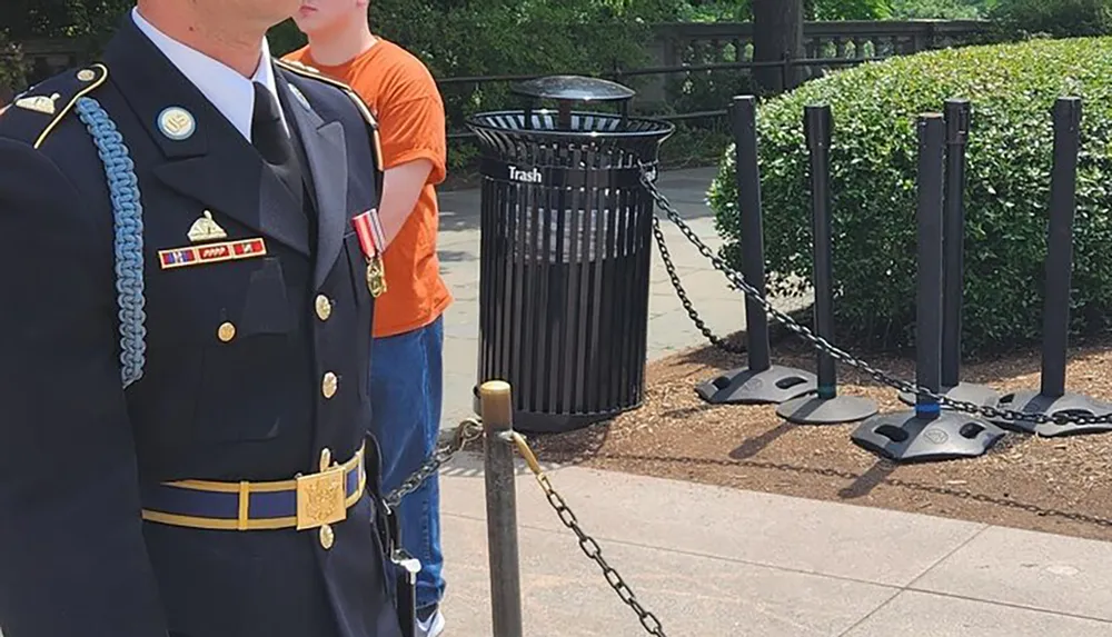 An individual in a military uniform stands near a young civilian on a sunny day with a trash receptacle and a chain-link barrier partially visible in the background