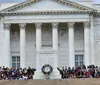 The image shows a group of people sitting on the steps of a neoclassical building with columns and a wreath possibly during a public event or ceremony