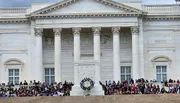 The image shows a group of people sitting on the steps of a neoclassical building with columns and a wreath, possibly during a public event or ceremony.