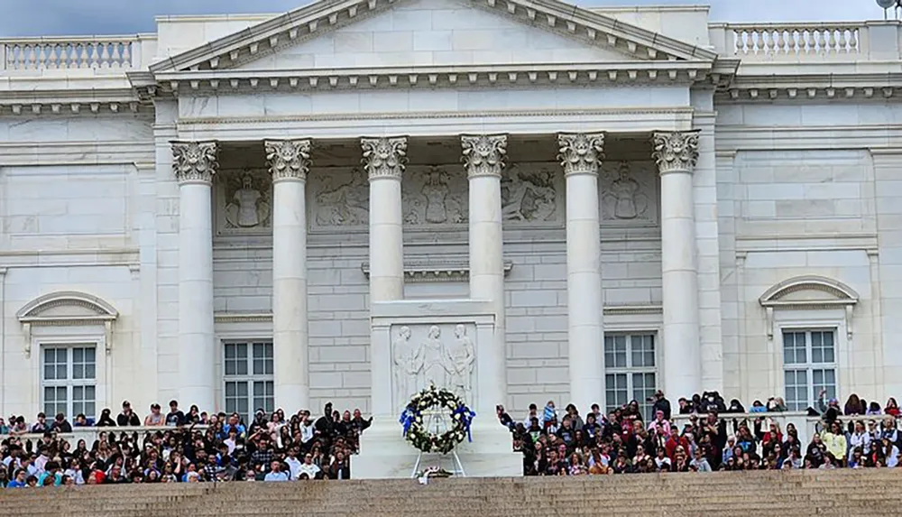 The image shows a group of people sitting on the steps of a neoclassical building with columns and a wreath possibly during a public event or ceremony