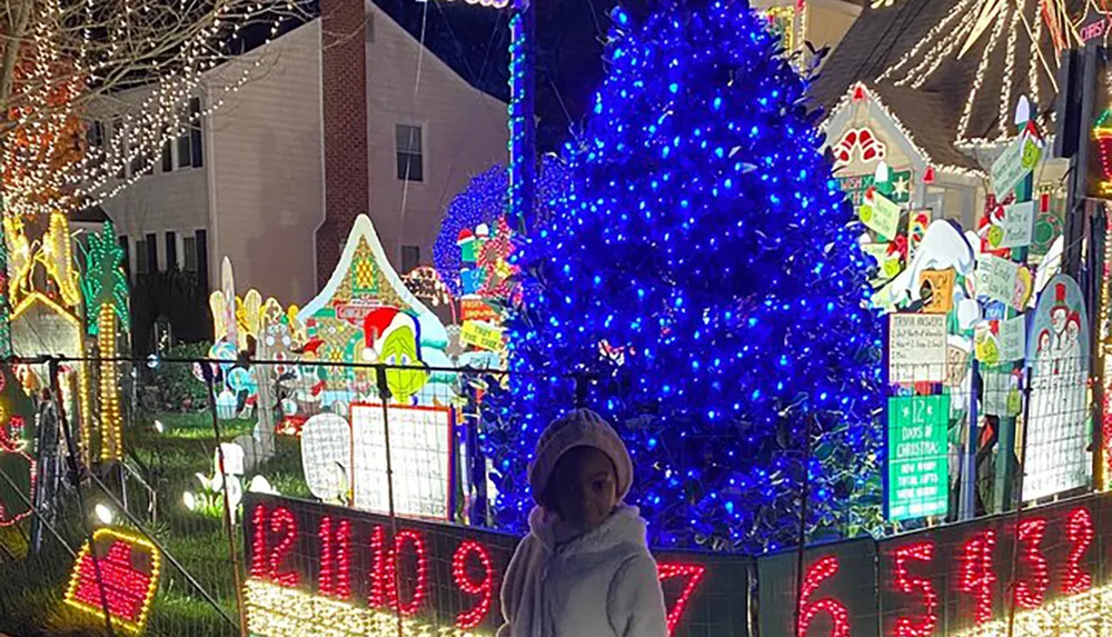 A person stands in front of a house extravagantly decorated with vibrant Christmas lights and festive displays