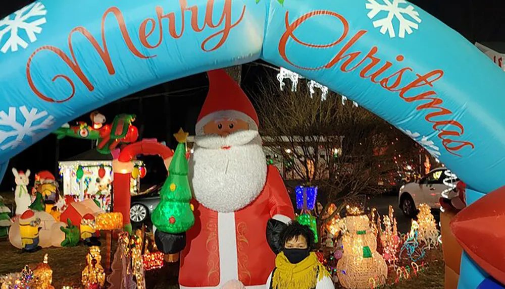 A vibrant outdoor Christmas display featuring an inflatable Santa Claus a colorful arch reading Merry Christmas and various illuminated decorations with a person standing in front