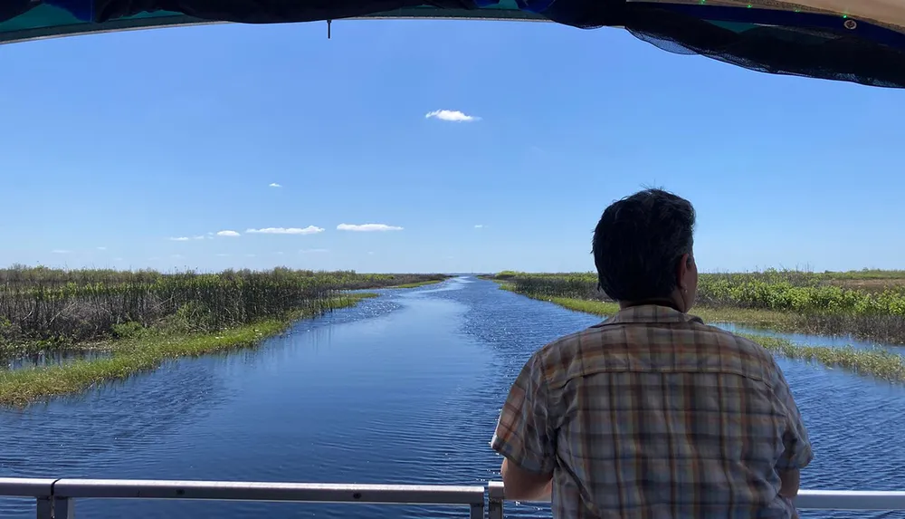 A person is gazing at a serene waterway surrounded by wetlands under a clear blue sky