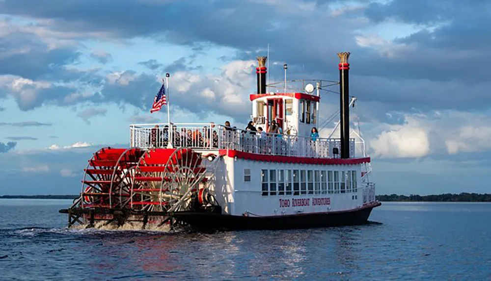 The image shows a red and white paddle steamer with passengers on board adorned with an American flag cruising on a body of water under a partly cloudy sky