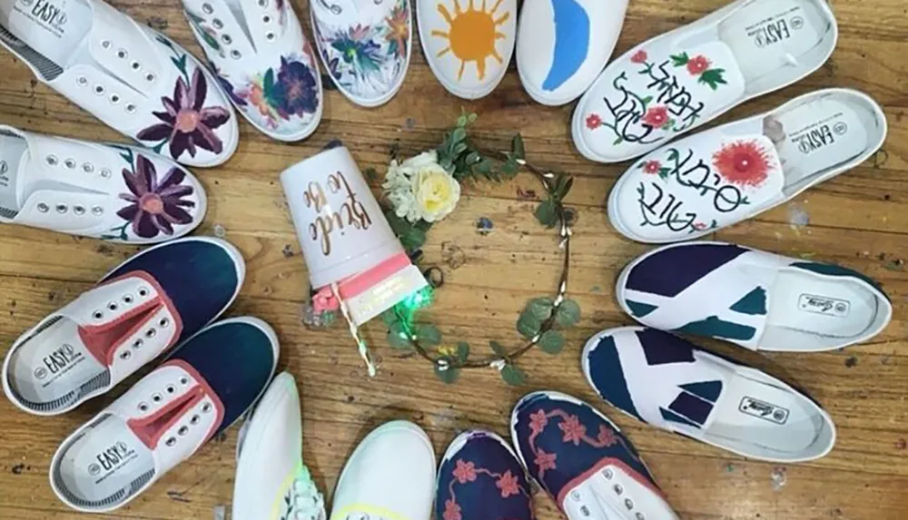 The image shows a variety of shoes artistically arranged in a circle each pair adorned with different colorful designs and a decorated tumbler and flower arrangement are placed at the center of the circle