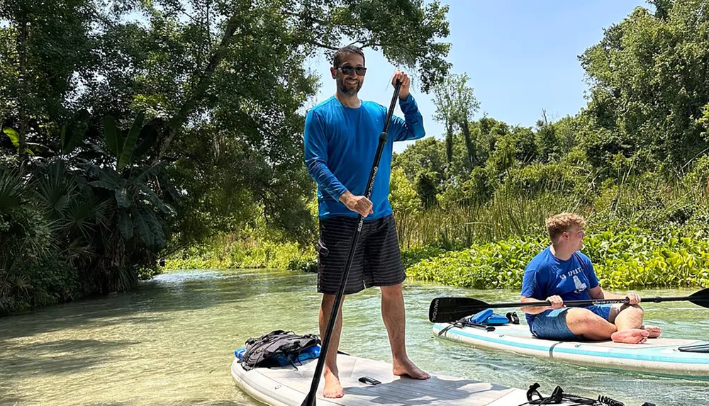 Two people are enjoying paddleboarding on a calm river surrounded by lush greenery on a sunny day