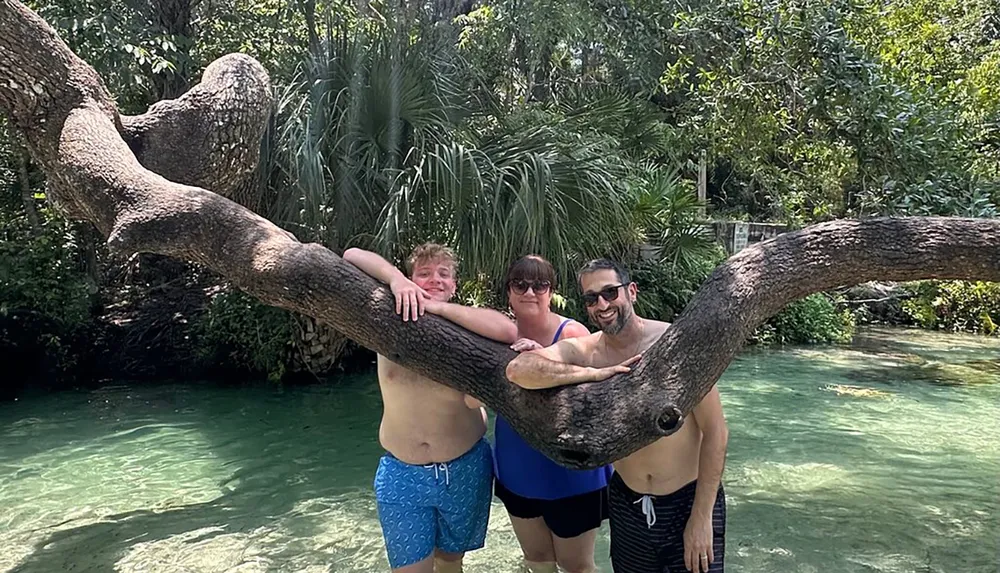 Three people are smiling and posing with their arms draped over a large tree branch by a clear water stream in a tropical environment