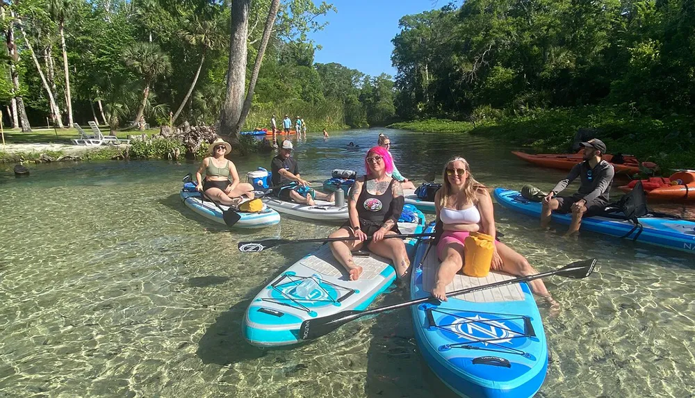 A group of people are enjoying a sunny day on a clear water river with stand-up paddleboards and kayaks surrounded by lush greenery