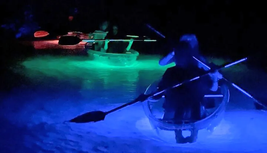 People are kayaking at night with the water illuminated by colorful lights beneath them.