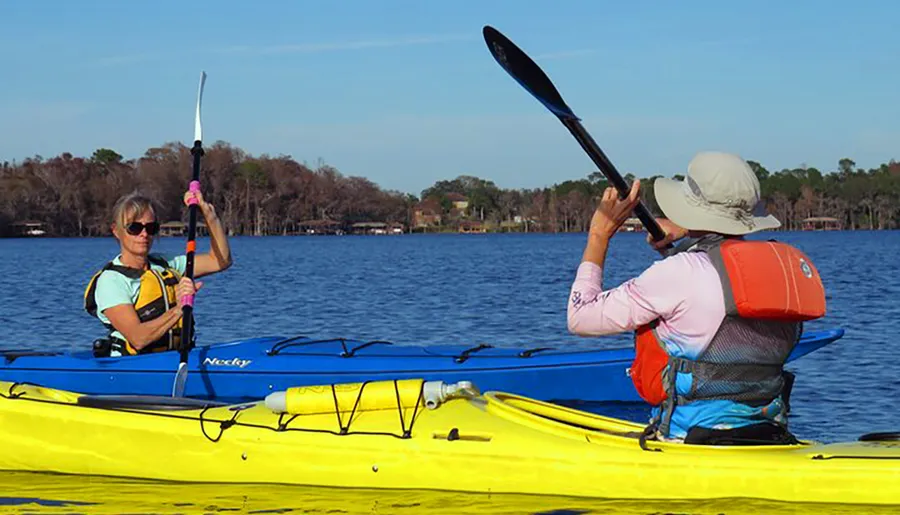 Two people are kayaking on a serene blue lake under a clear sky.