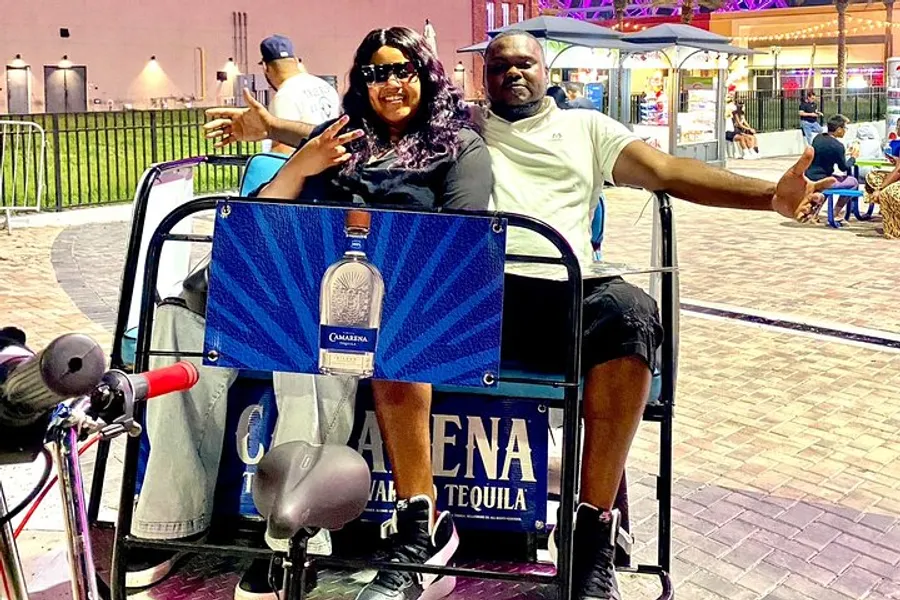 A smiling couple is enjoying a ride on a pedicab adorned with a tequila advertisement in an outdoor setting during the evening.