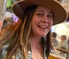 A smiling person with a hat gives a peace sign while holding a glass of beer in a lively indoor setting