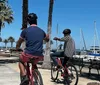 Two individuals are riding bicycles by a marina lined with palm trees and sailboats on a sunny day