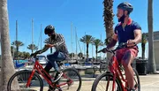 Two individuals are riding bicycles by a marina lined with palm trees and sailboats on a sunny day.