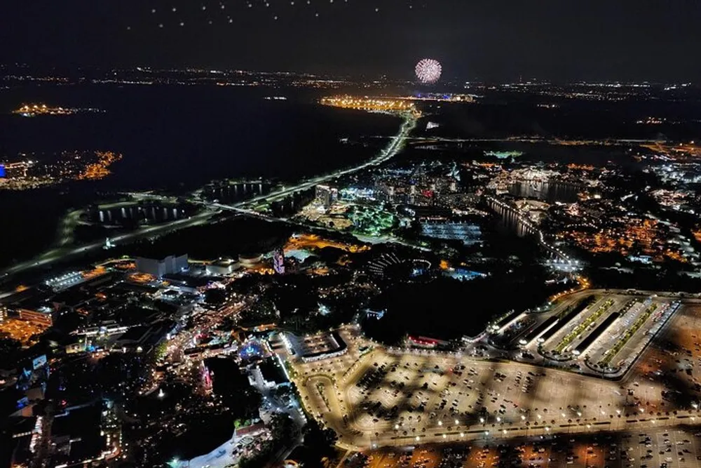 The image shows a nighttime aerial view of a vibrant illuminated cityscape with a bridge crossing a body of water and fireworks bursting in the distance