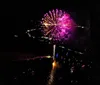 The image shows a nighttime aerial view of a vibrant illuminated cityscape with a bridge crossing a body of water and fireworks bursting in the distance