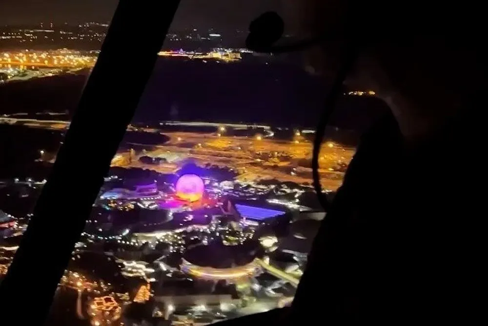 The image appears to be taken from the cockpit of an aircraft during nighttime showing a well-lit and colorful urban area with a prominent illuminated round structure below as seen through the aircrafts windows with part of the pilots silhouette visible