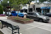 The photo captures a peaceful street view, showcasing parked cars, a vibrant blue Jeep, a sitting area with a bench, and a tree-lined sidewalk in an urban setting.