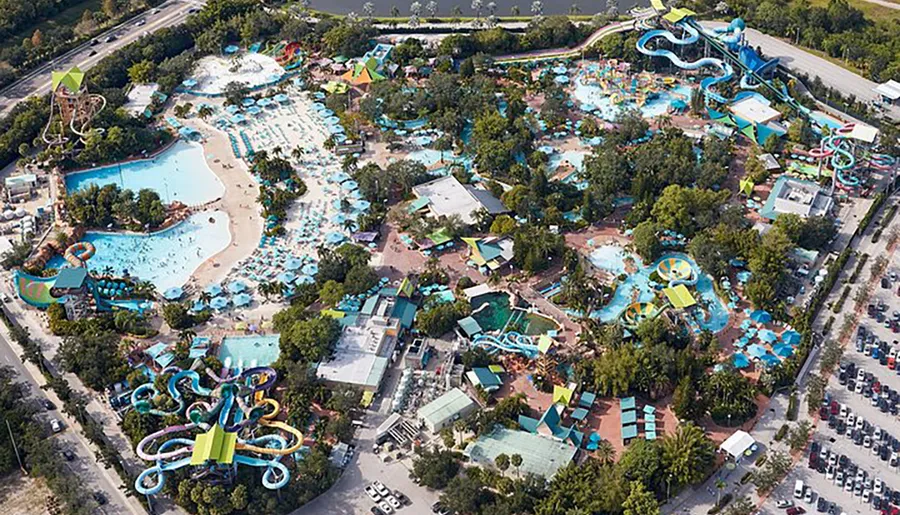 The image shows an aerial view of a colorful water park with a variety of slides, pools, and leisure areas surrounded by parking and greenery.