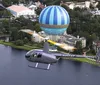 A helicopter is depicted flying close to a large anchored hot air balloon over a scenic area with buildings and a body of water