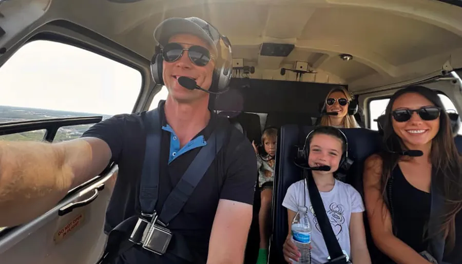 A pilot is taking a selfie with four passengers, all wearing headsets, in a helicopter cockpit.