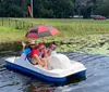 Three people are enjoying a sunny day on a pedal boat in a pond with lily pads while one of them holds an open umbrella for shade