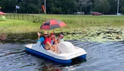 Three people are enjoying a sunny day on a pedal boat in a pond with lily pads, while one of them holds an open umbrella for shade.
