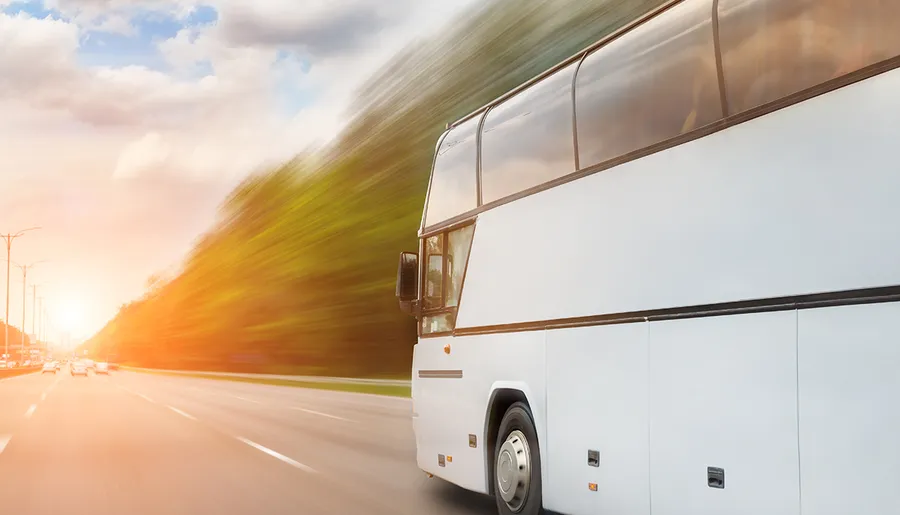 A white coach bus is shown in motion on a road with blurred scenery indicating high speed, and the bright sun visible in the background, suggesting either dawn or dusk.